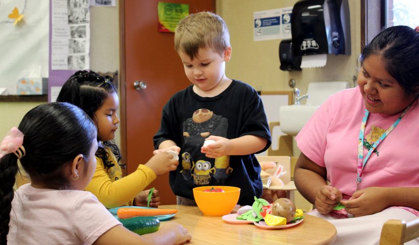 Three students and a teaching assistant sitting around a table doing imaginary play with toy foods